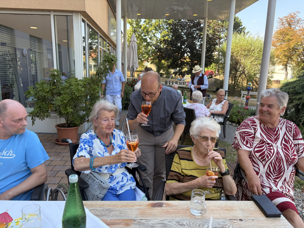 ein Herr und zwei ältere Damen mit einem Glas Aperol und ein junger Mann trinkt vom Aperol