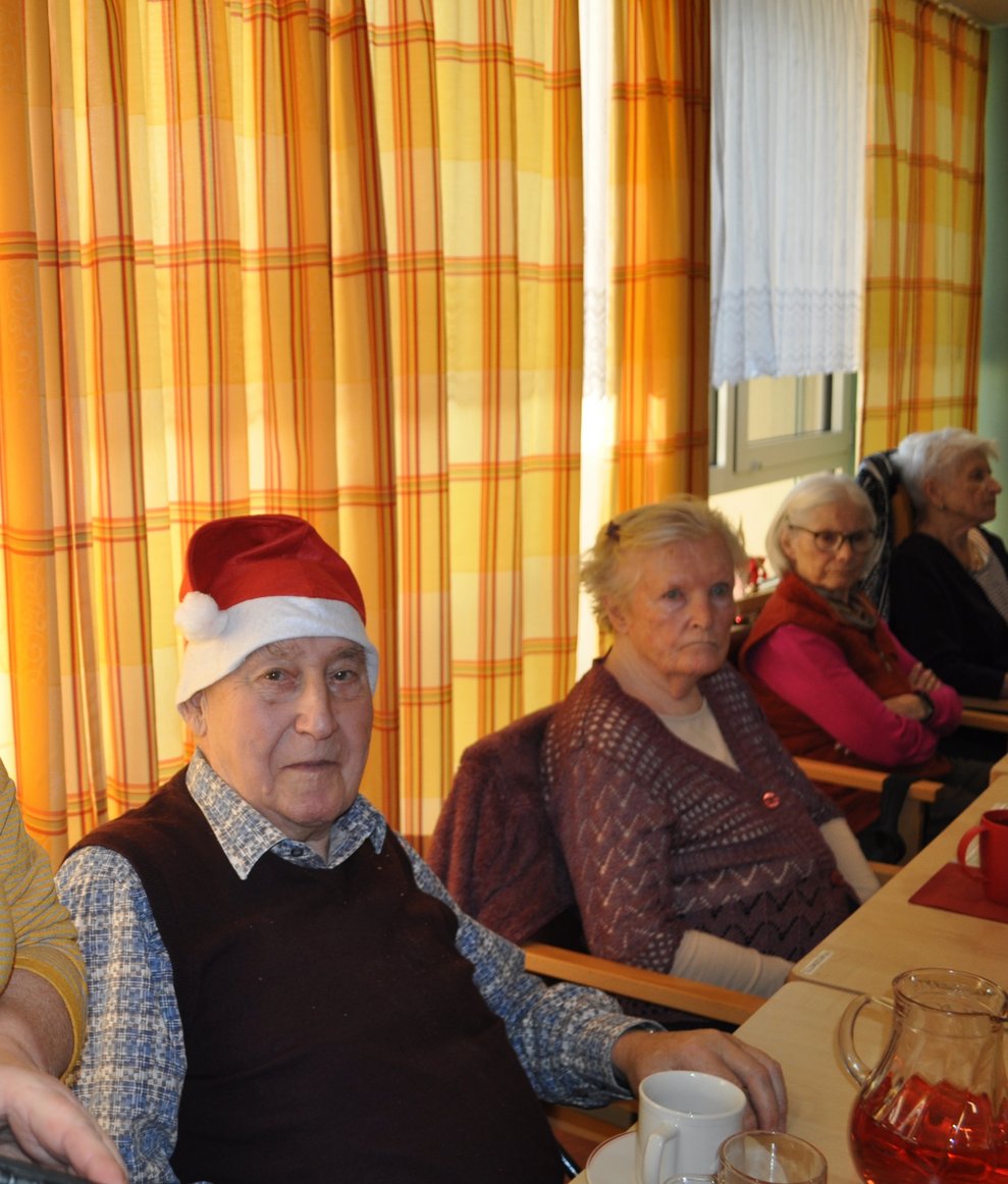 ein älterer Herr mit Weihnachtsmann Mützen sowie weiteren älteren Personen und weihnachtlich gedeckten Tischen