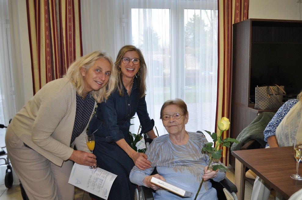 eine ältere Dame bekommt Pralinen und eine Blume geschenkt von zwei Damen