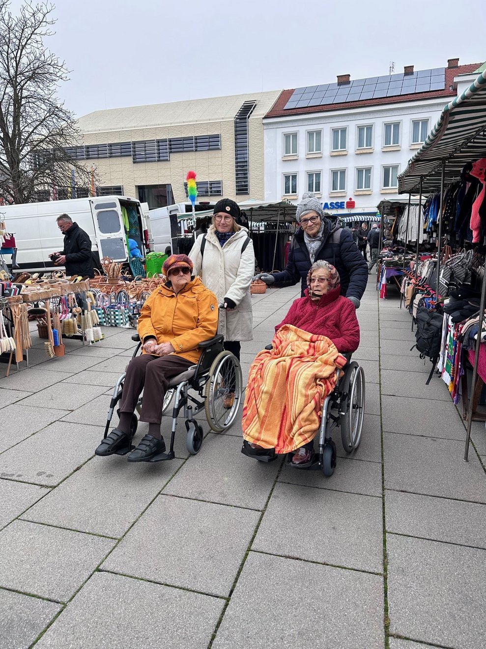zwei Damen im Rollstuhl mit einer Begleitperson am Markt