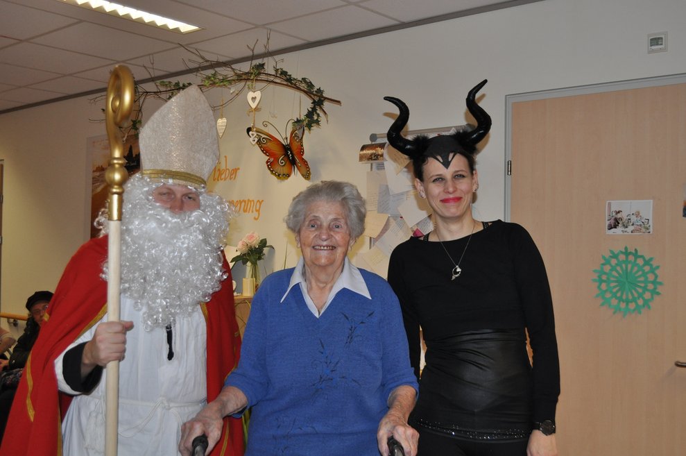 eine Frau als Krampus verkleidet und ein Mann als Nikolaus verkleidet zu Besuch bei einer älteren Dame im Pflegeheim