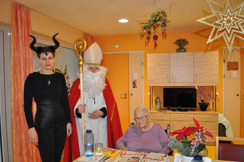 eine Frau als Krampus verkleidet und ein Mann als Nikolaus verkleidet zu Besuch bei einer älteren Dame im Pflegeheim