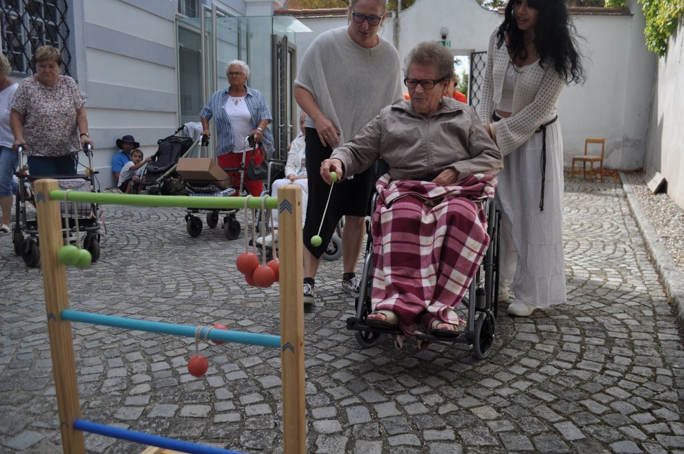 eine ältere Dame im Rollstuhl versucht sich an einem Stand mit einem lustigen Wurfspiel