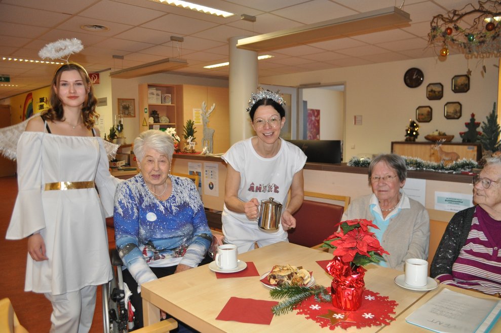 eine junge Frau als Engel verkleidet, drei ältere Damen bei einem weihnachtlich dekorierten Tisch und eine Mitarbeiterin schenkt Tee aus einer Kanne aus