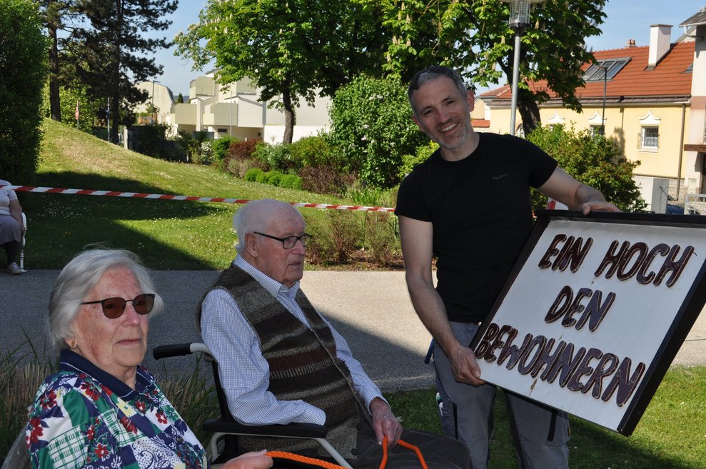 eine ältere Dame und ein älterer Herr mit einem Schild für den Maibaum wo steht "ein Hoch den Bewohnern"