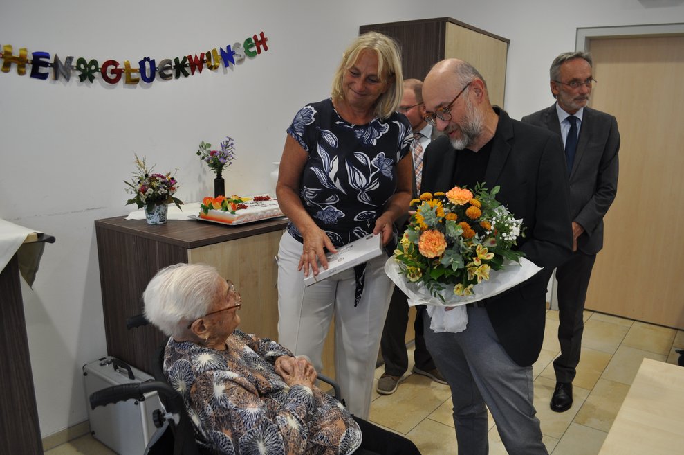 ein Mann mit einem Blumenstrauß und eine Frau mit Pralinen gratulieren einer älteren Dame zum 100. Geburtstag 
