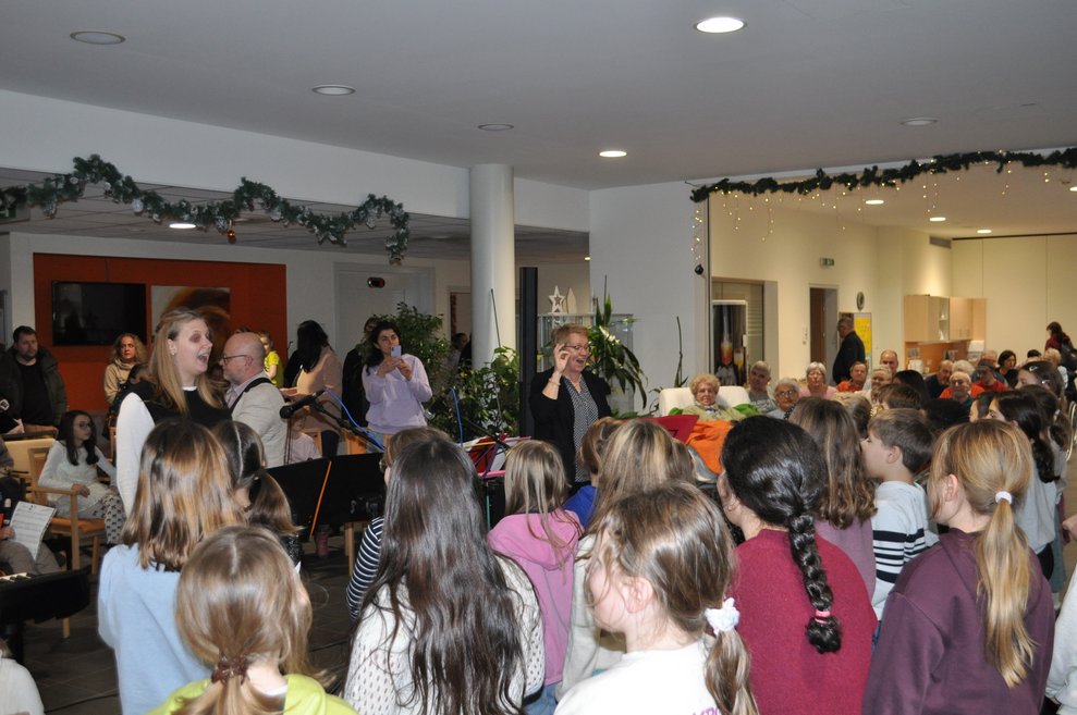 einige Kinder singen im Chor, davor steht die Chorleiterin und dirigiert, dahinger einige ältere Menschen als Zuseher