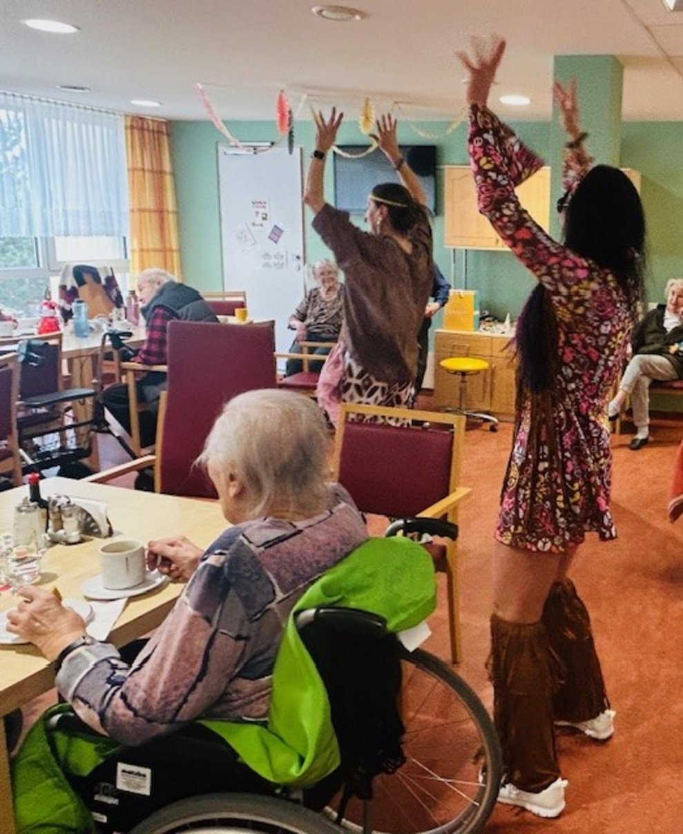 einige Menschen sitzen bei Tisch und zwei Frauen sind als Hippies verkleidet und klatschen über dem Kopf in die Hände