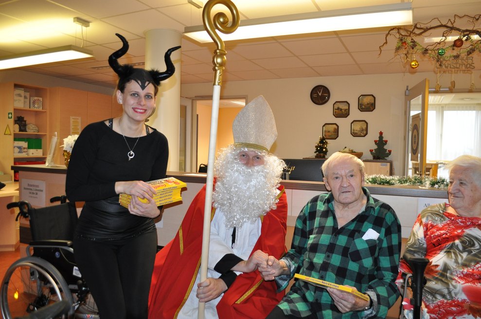 eine Frau als Krampus verkleidet und ein Mann als Nikolaus verkleidet zu Besuch bei einer älteren Dame und einem älteren Herrn im Pflegeheim