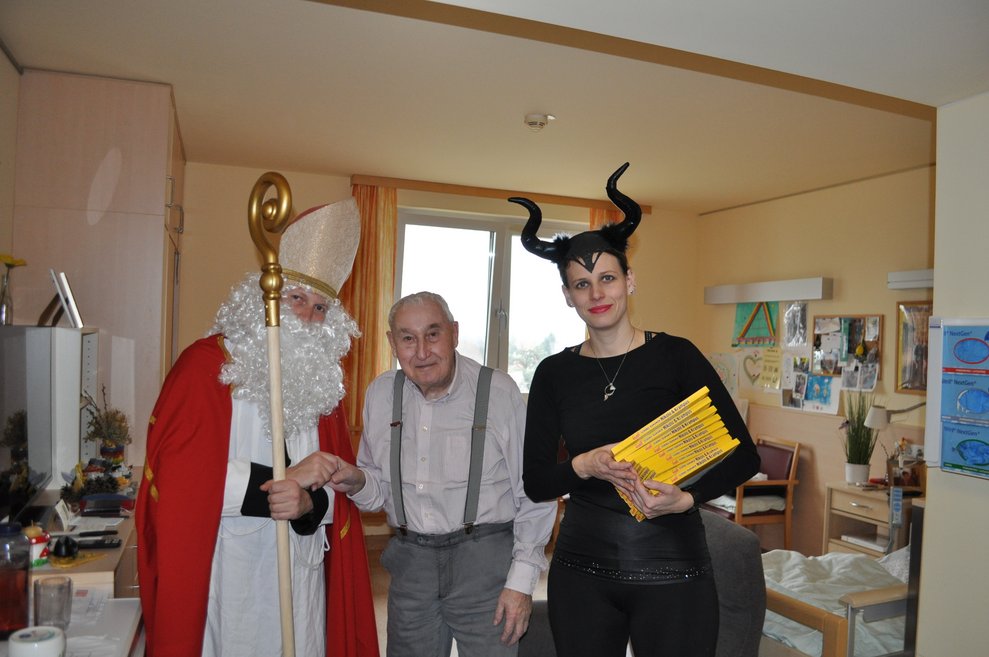 eine Frau als Krampus verkleidet und ein Mann als Nikolaus verkleidet zu Besuch bei einem Herrn im Pflegeheim