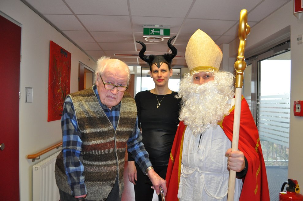 eine Frau als Krampus verkleidet und ein Mann als Nikolaus verkleidet zu Besuch bei einem Herrn im Pflegeheim
