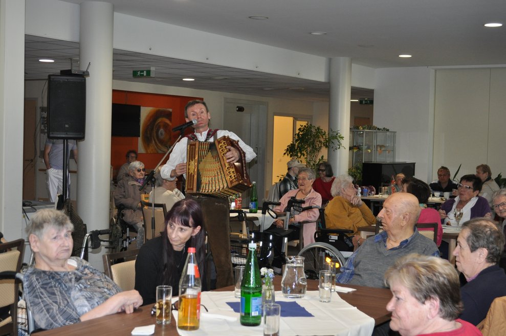 einige Menschen sitzen bei Tisch und hören dem Ziehharmonika Spieler zu