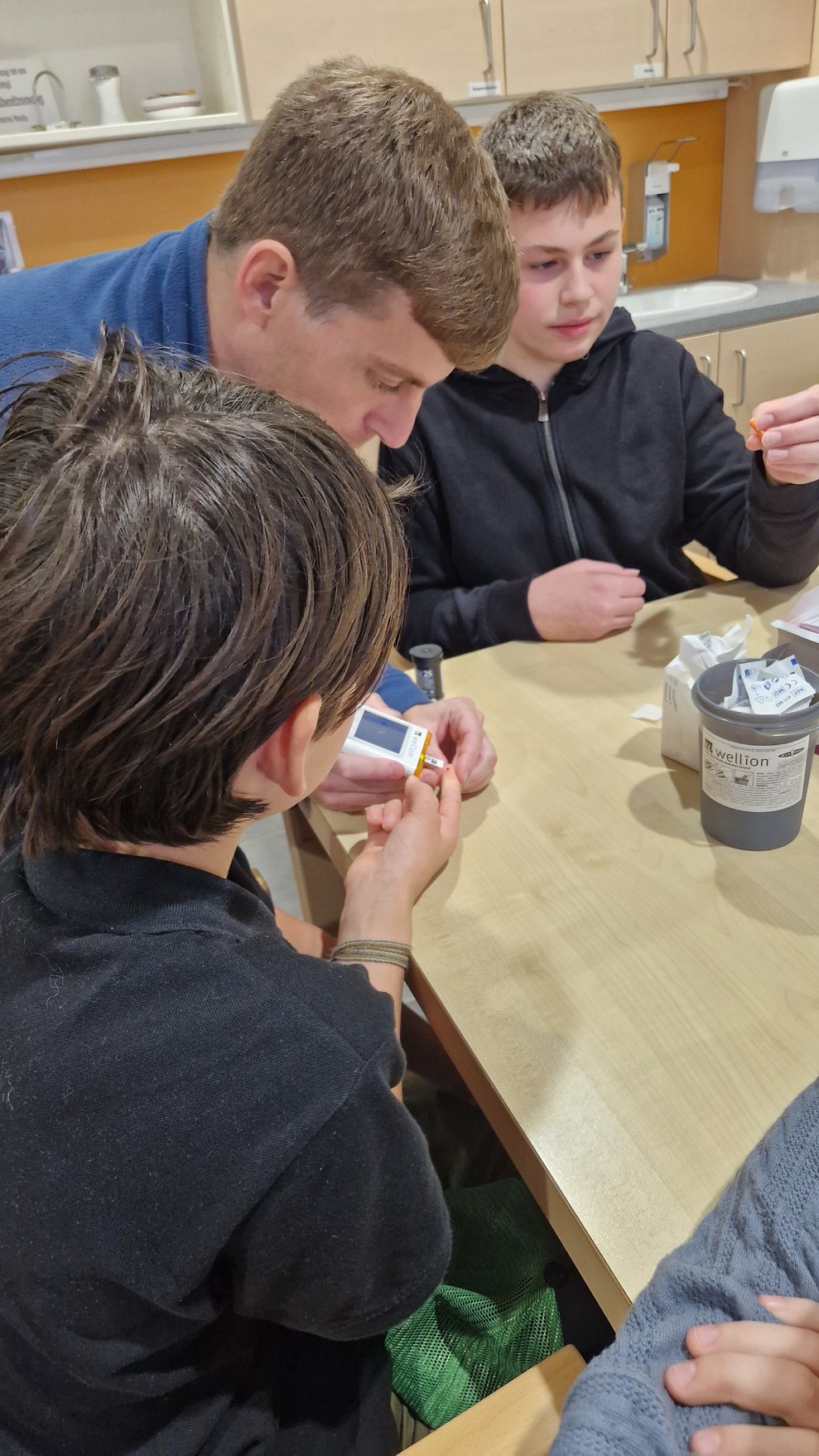 ein Mann zeigt zwei Buben wie man Blutzucker misst