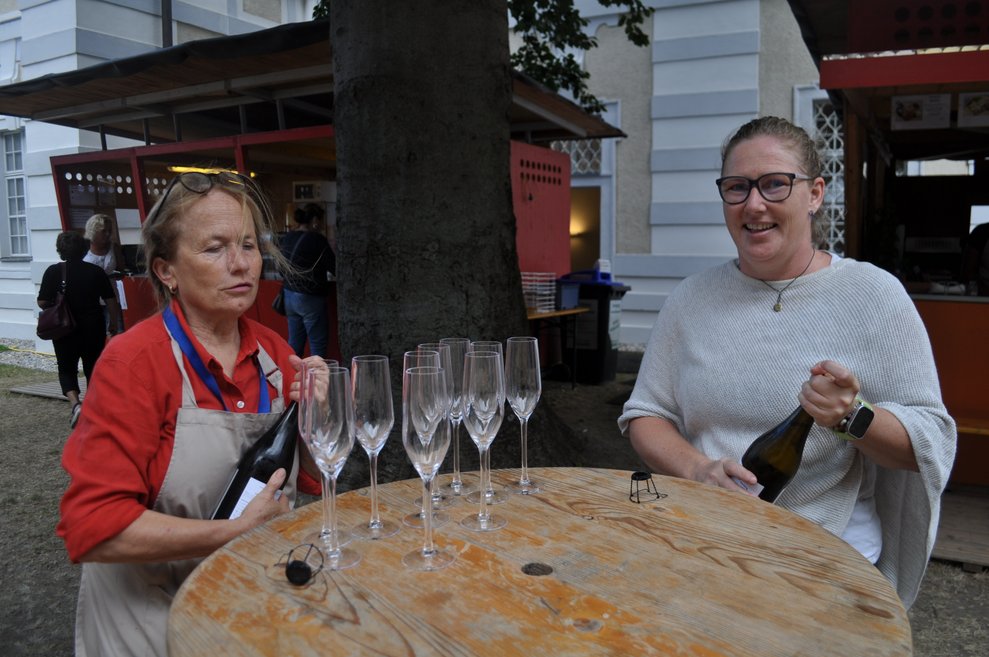 Eine Dame mit Schürze schenkt Sekt in Gläser, eine zweite Frau hält eine Sektflasche in den Händen