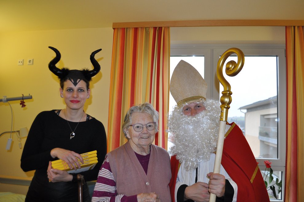 eine Frau als Krampus verkleidet und ein Mann als Nikolaus verkleidet zu Besuch bei einer älteren Dame im Pflegeheim