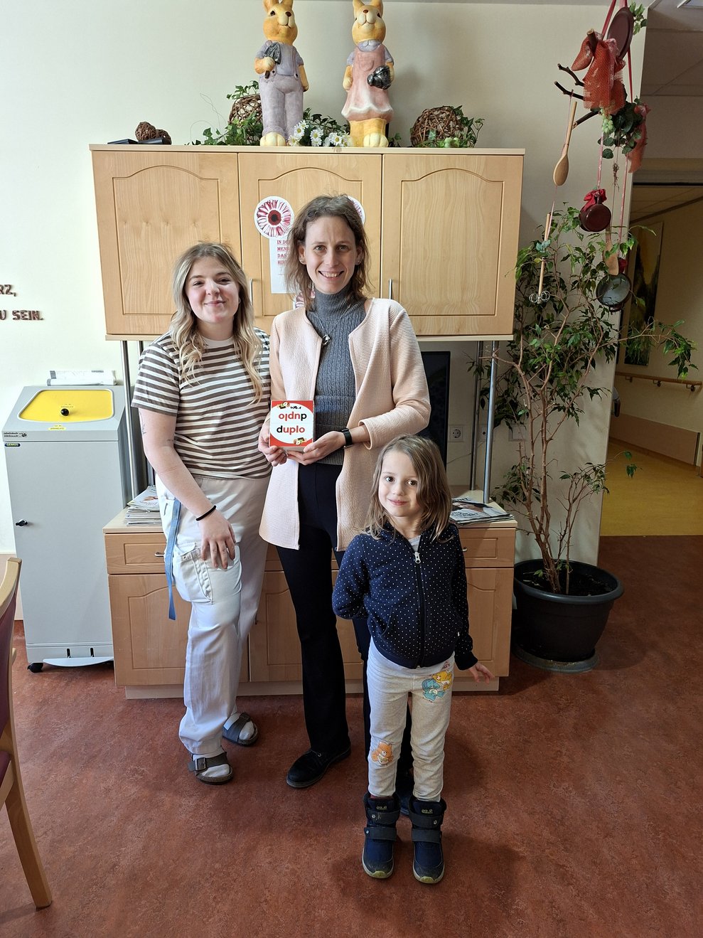 2 Mitarbeiterinnen und 1 Mädchen im Wohnbereich bei der Übergabe der Duplo Riegel.