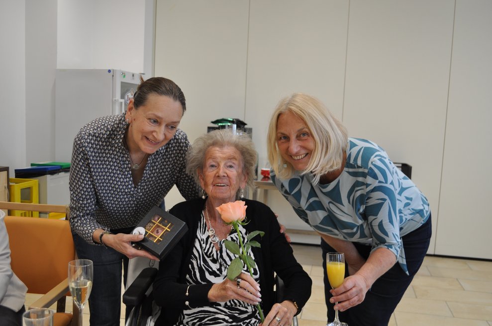 zwei Frauen gratulieren einer älteren Dame zum Geburtstag und überreichen Pralinen und eine Rose