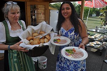 Eine Frau hält einen Korb voll Semmeln und eine zweite Frau hält zwei Teller mit saurer Wurst und Garnierung