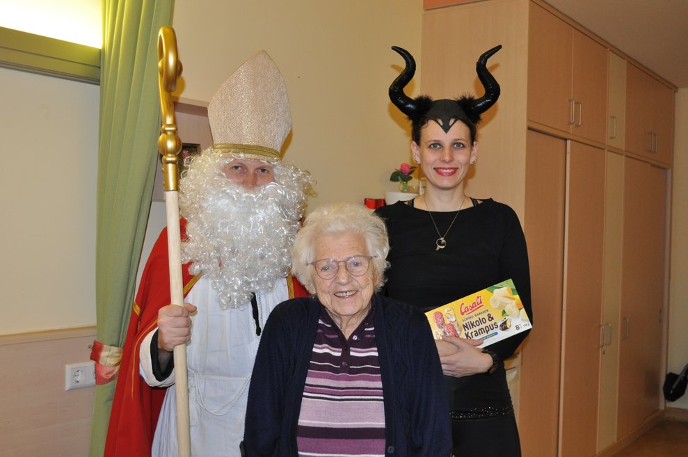 eine Frau als Krampus verkleidet und ein Mann als Nikolaus verkleidet zu Besuch bei einer älteren Dame im Pflegeheim