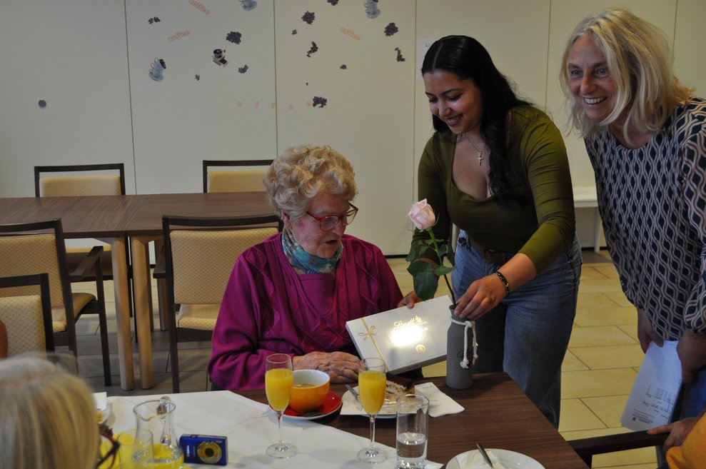 eine ältere Dame bekommt Pralinen und eine Blume geschenkt von zwei Damen