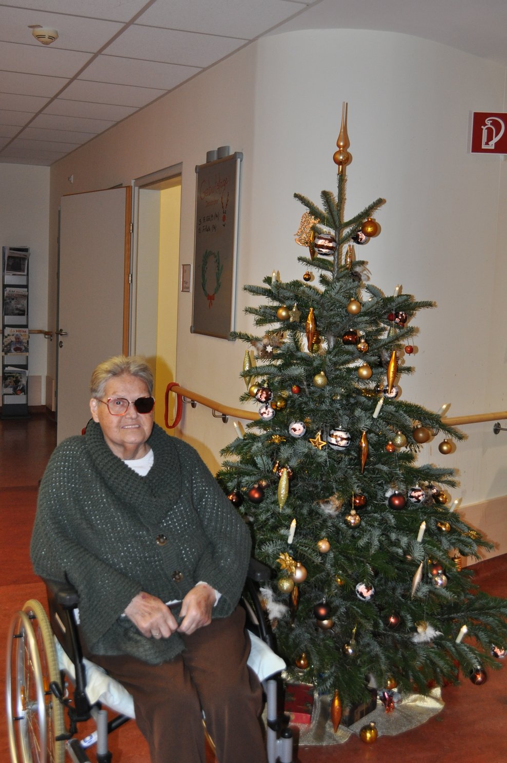 eine ältere Dame sitzt im Rollstuhl vor einem geschmückten Christbaum