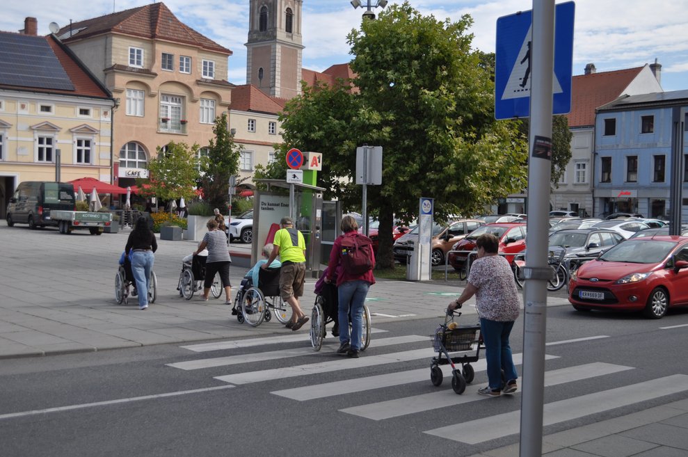 Mehrere Personen in Rollstühlen und Rollatoren überqueren eine Straße auf einem Zebrastreifen