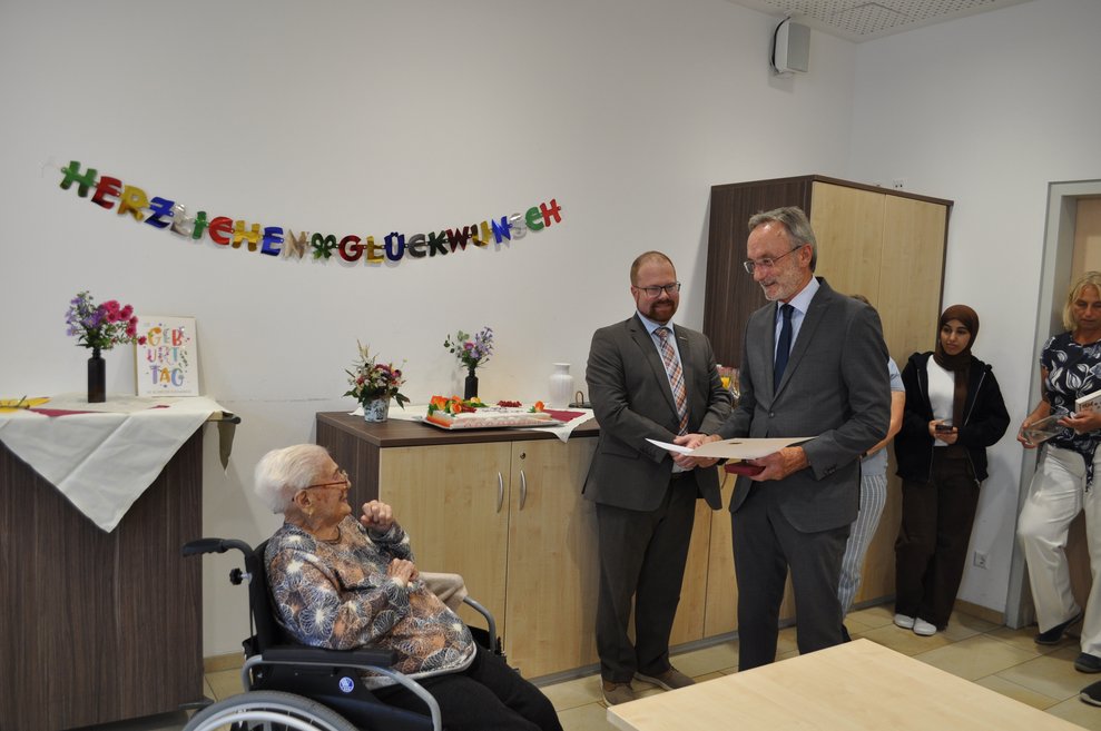 ein Mann mit einer Urkunde der einer älteren Dame zum 100. Geburtstag gratuliert