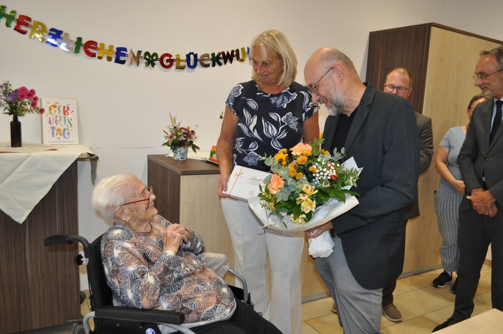 ein Mann mit einem Blumenstrauß und eine Frau mit Pralinen gratulieren einer älteren Dame zum 100. Geburtstag 