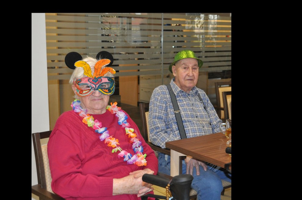 eine ältere Dame mit Maske und Hawai Kette und ein älterer Herr mit grünem Hut