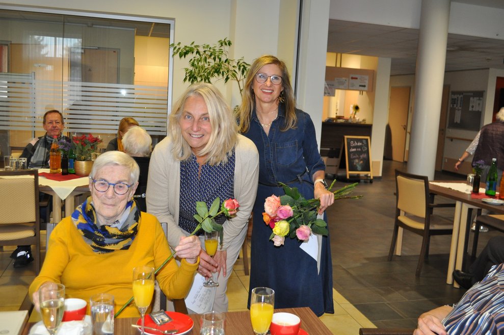 eine ältere Dame bekommt Pralinen und eine Blume geschenkt von zwei Damen