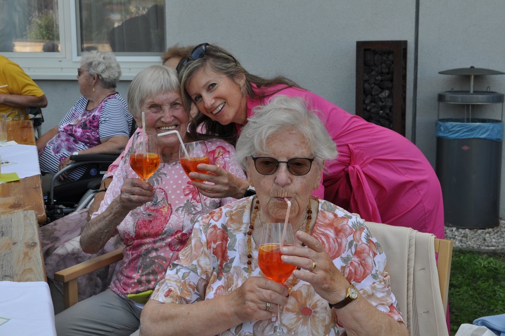 zwei ältere Damen und eine Mitarbeiterin stoßen mit Aperol an