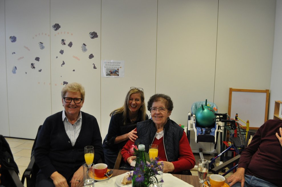 eine ältere Dame bekommt Pralinen und eine Blume geschenkt von zwei Damen