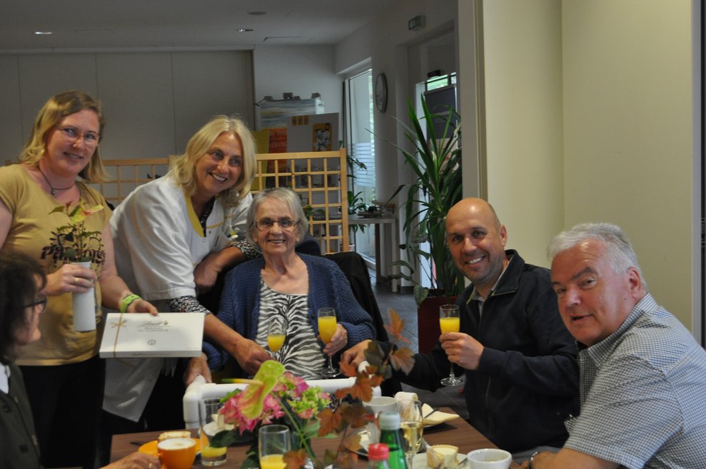 einige Menschen bei einer Geburtstagsfeier mit Blumen, Sekt und Pralinen