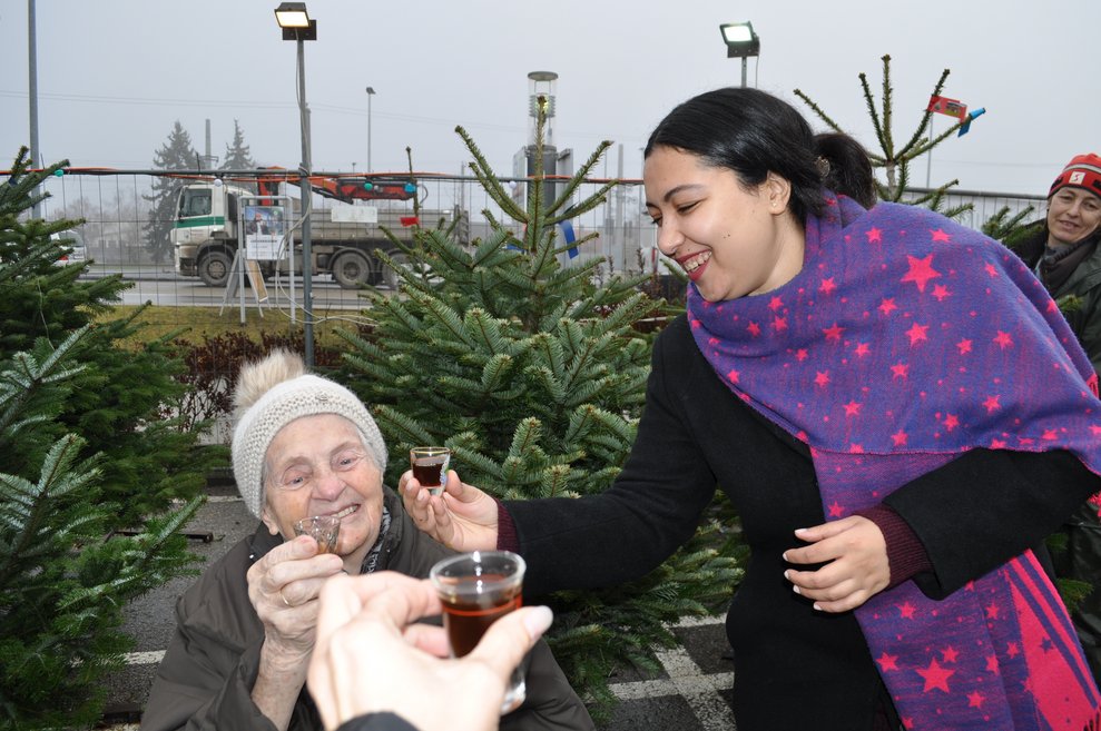 eine ältere und eine jüngere Damen stoßen mit einem kleinen Getränk an beim Christbaumverkaufsstand