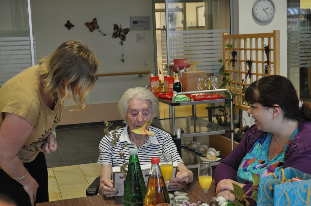 eine ältere Dame und zwei jüngere Damen beim Geburtstagsfest mit Blumen, Sekt und Kuchen