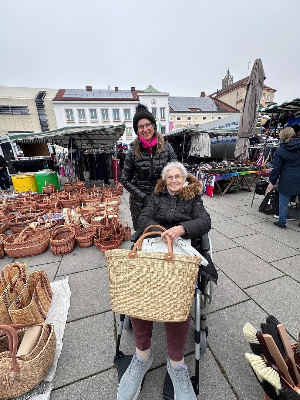 eine Dame im Rollstuhl mit einem großen Korb und mit einer Begleitperson am Markt