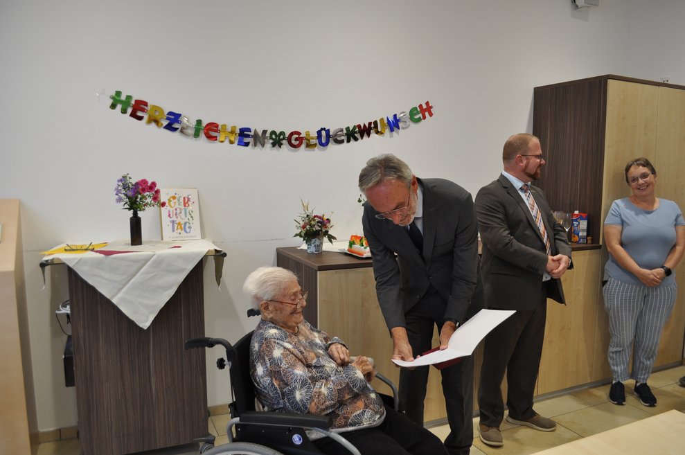 ein Mann mit einer Urkunde der einer älteren Dame zum 100. Geburtstag gratuliert