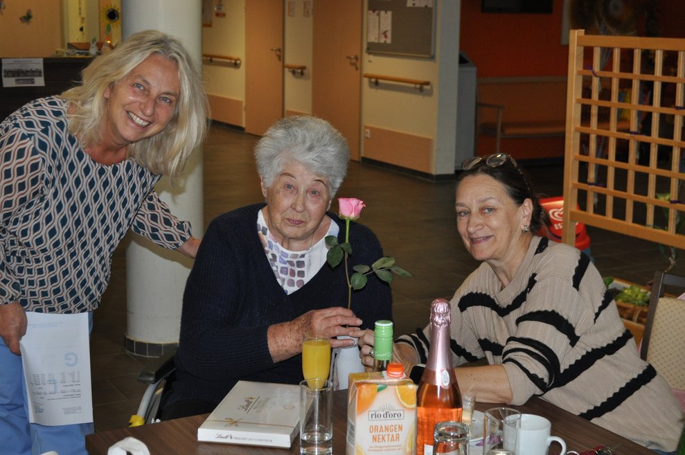 eine ältere Dame mit Sekt und Blumen bei der Geburtstagsfeier mit zwei anderen Frauen