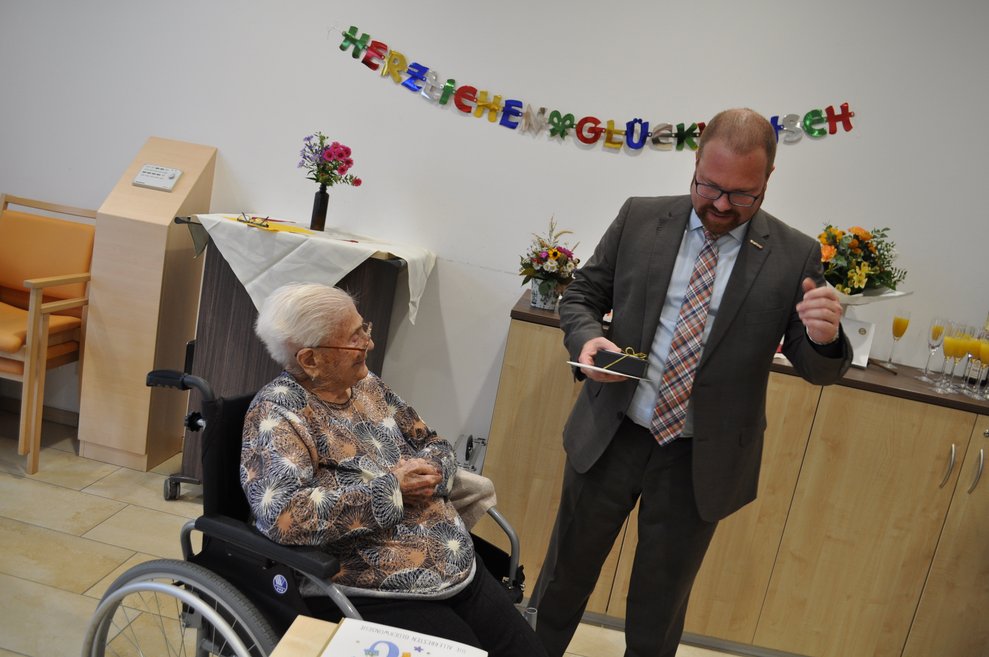 ein Mann mit einem kleinen Geschenk, der einer älteren Dame zum 100. Geburtstag gratuliert